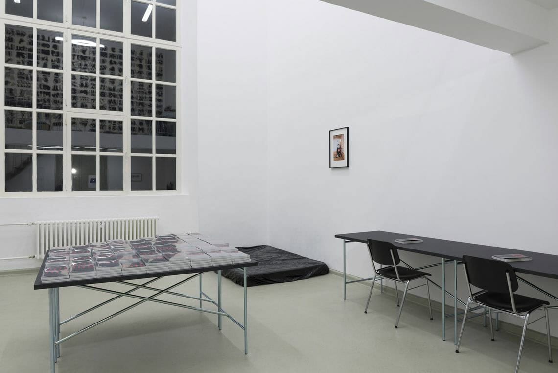 Foto: Ein Raum mit weißen Wänden und grauem Boden. Im Raum stehen zwei Tische mit schwarzen Tischplatten. Einer der tische ist eingeschrägt. auf ihm liegen Stapel aus Heften, die jeweils in Folie eingeschlagen sind. An dem anderen Tisch stehen zwei schwarze Stühle, vor denen auf dem Tisch ebenfalls zwei der hefte liegen. In einer der Raumecken liegt eine schwarze Matte auf dem Boden. An der Wand hängt ein gerahmtes Objekt.