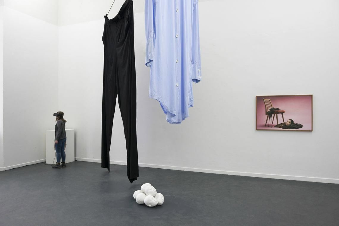 Foto: Ein Raum mit weißen Wänden und grauem Boden. Von der Decke Gängen Textilien. Auf dem Boden liegt eine Gruppe von Objekten, an der Wand hängt ein flaches Objekt. Im Raum steht eine Person die eine VR-Brille trägt.