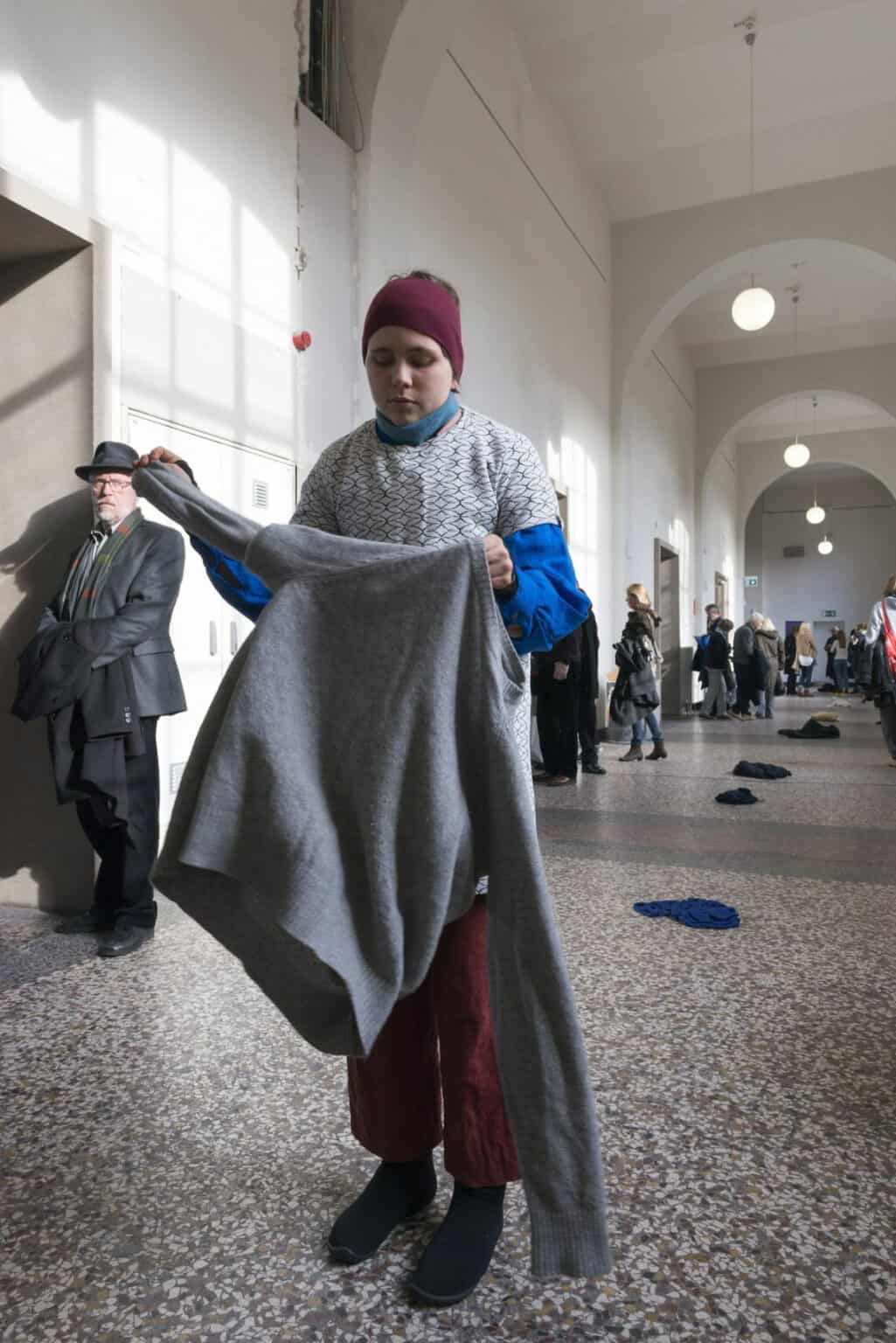 Foto: Ein langer Flur mit weißen Wänden und mehrfarbigem Steinboden. Im Flur befinden sich viele Menschen. Auf dem Boden liegen, in einer reihe hintereinander, mehrere Textilien auf dem Boden.