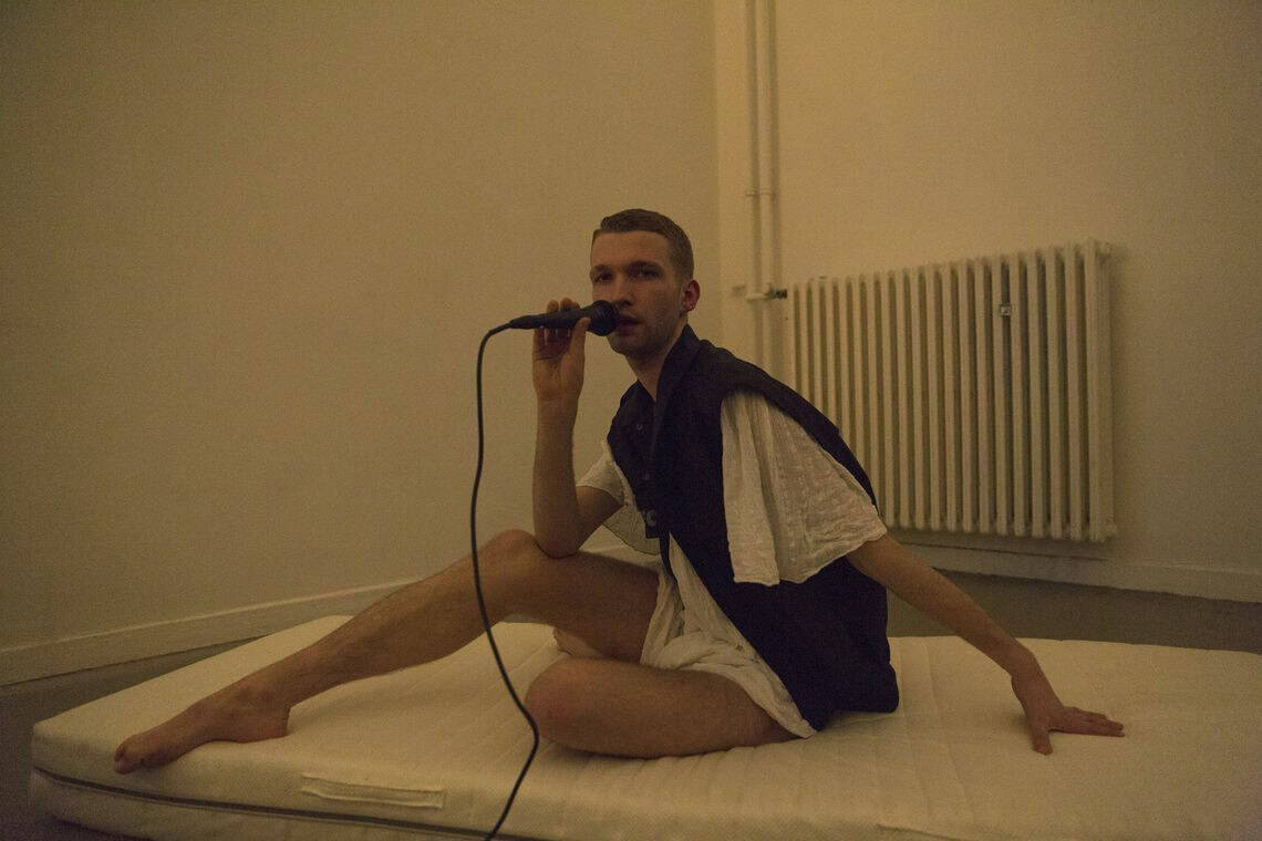 Foto: Eine ungezogene Matratze in einem Raum mit weißen Wänden. Auf der Matratze sitzt ein Mensch mit freigelegten Beinen und nach hinten gestreckten Arm. Er hält ein Mikrofon in der Hand und blickt in die Kamera.