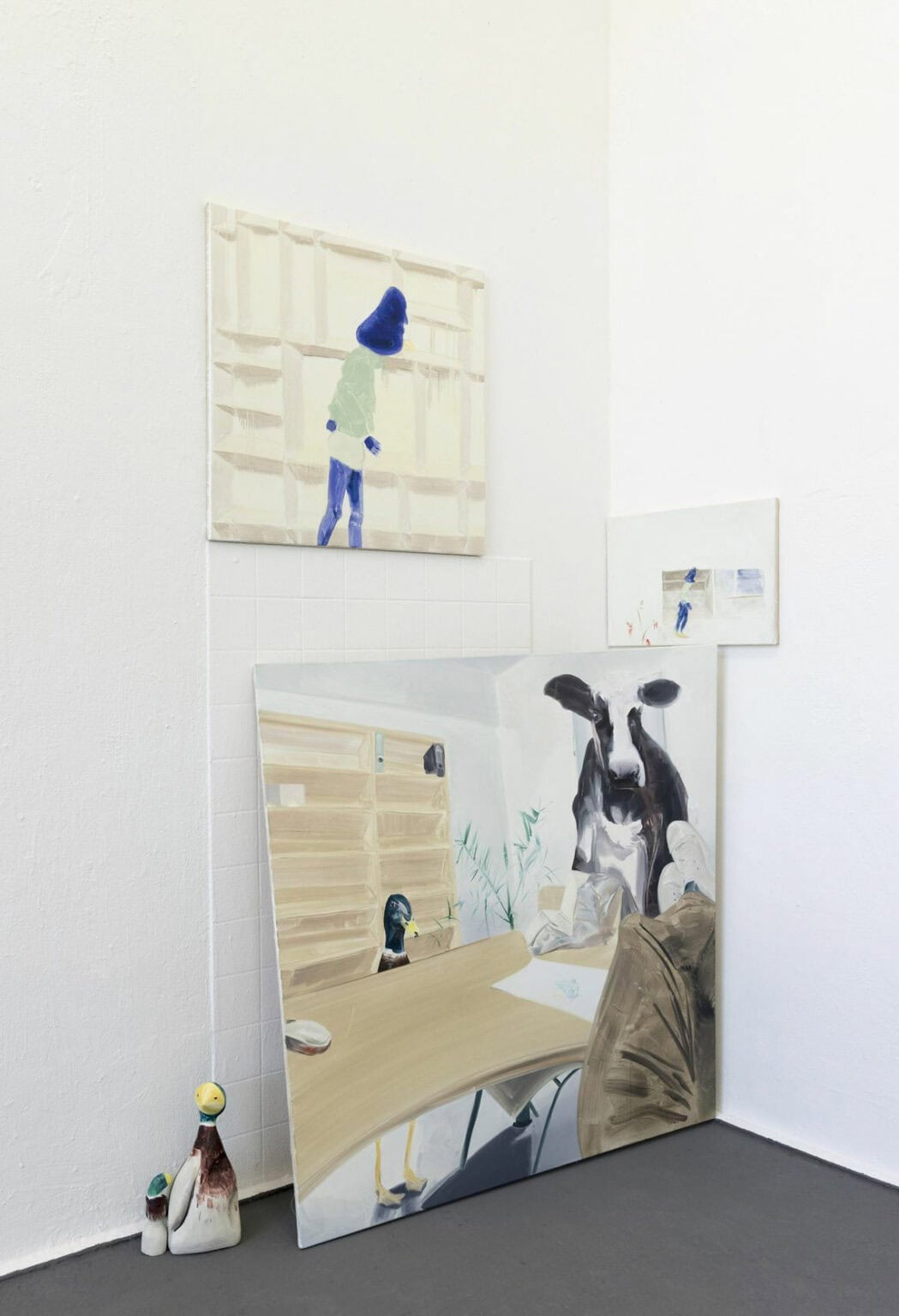 Foto: Eine Raumecke mit weißen Wänden und grauem Boden. An der Wand hängen und lehnen klein-, mittel- und großformatige Leinwände. Auf dem Boden Stehen zwei Objekte.