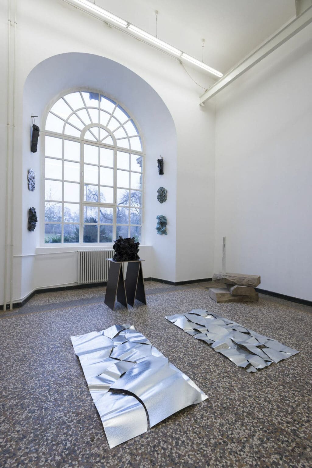 Foto: Ein Raum mit weißen Wänden, mehrfarbigem Steinboden und einem tieferliegenden Bogenfenster. In der Einbuchtung um das Fenster herum befinden sich, übereinander hängend, mehrere Objekte. Auf dem Boden stehen und liegen weitere Skulpturen.