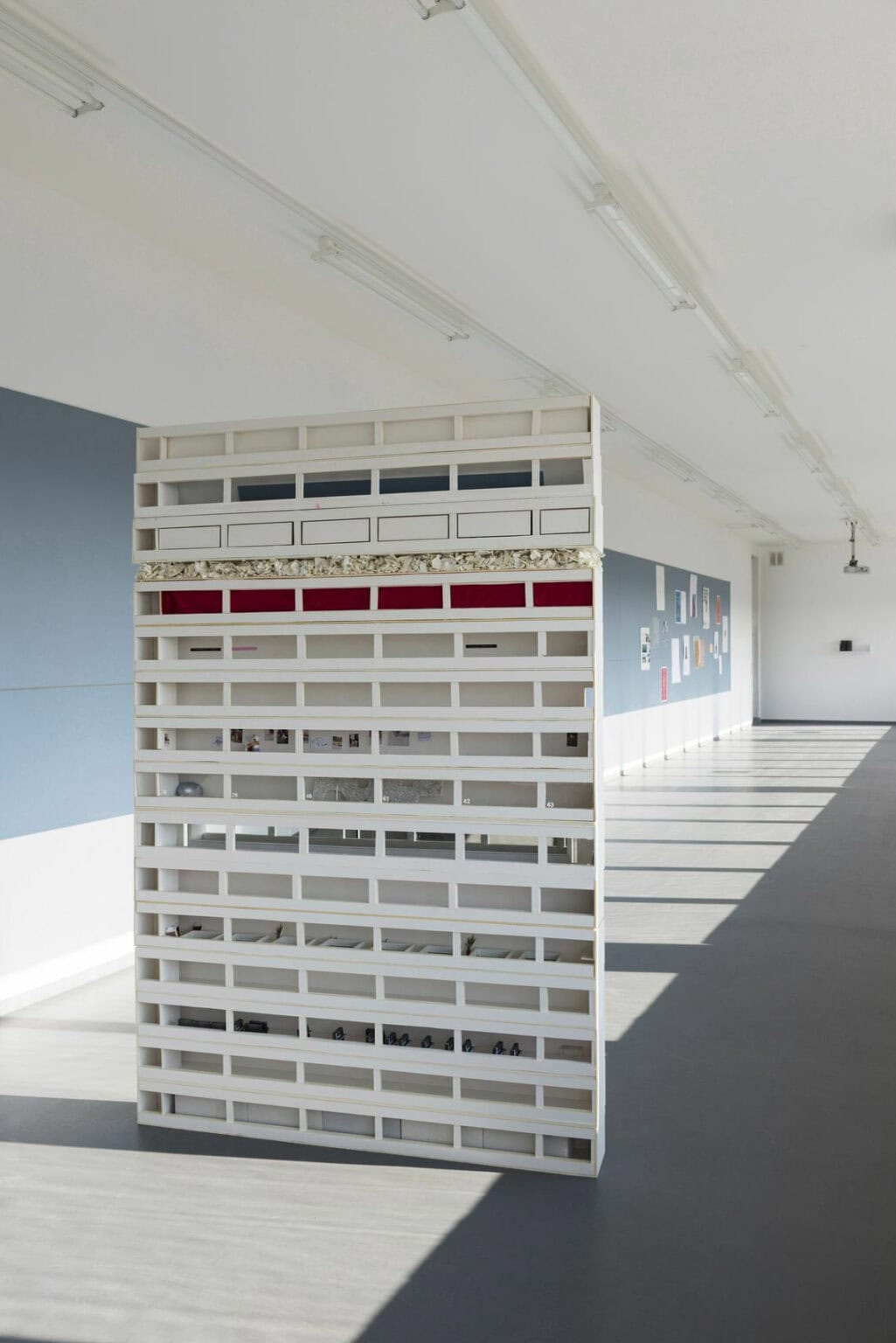 Foto: Ein schmaler, langer Raum mit weißen Wänden und grauem Boden. Auf die Wand ist ein hellblauer Streifen angebracht, auf dem stellenweise Papiere hängen. Im Raum steht ein hohes, rechteckiges Objekt auf unterschiedlichen Materialien.