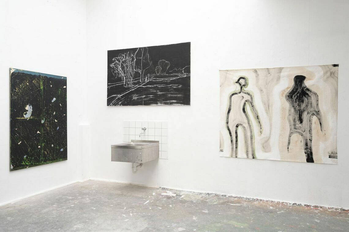 Foto: Ein Raum mit weißen Wänden und grauem Boden, der mit Farbflecken versehen ist. An den Wänden hängen, auf unterschiedlicher Höhe, großformatige Arbeiten auf Leinwand und Papier.