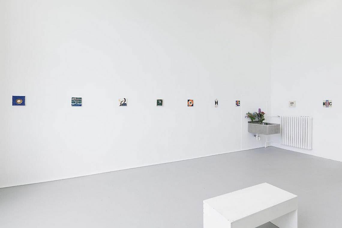 Foto: Ein Raum mit weißen Wänden, grauem Boden und einem Waschbecken. im Raum steht eine Sitzgelegenheit. An den Wänden hängen, auf einer Linie und im gleichen Abstand zueinander, kleinformatige Objekte.