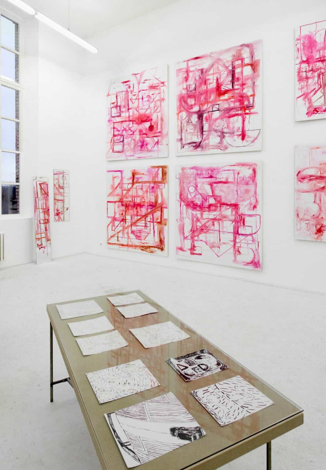 Foto: Ein Raum mit weißen Wänden und weißem Boden. An der Wand hängen mehrere großformatige Leinwände neben- und übereinander. Eine Leinwand lehnt an der Wand. Im Raum steht ein Tisch mit einer Glasplatte, auf dem Papierarbeiten liegen.