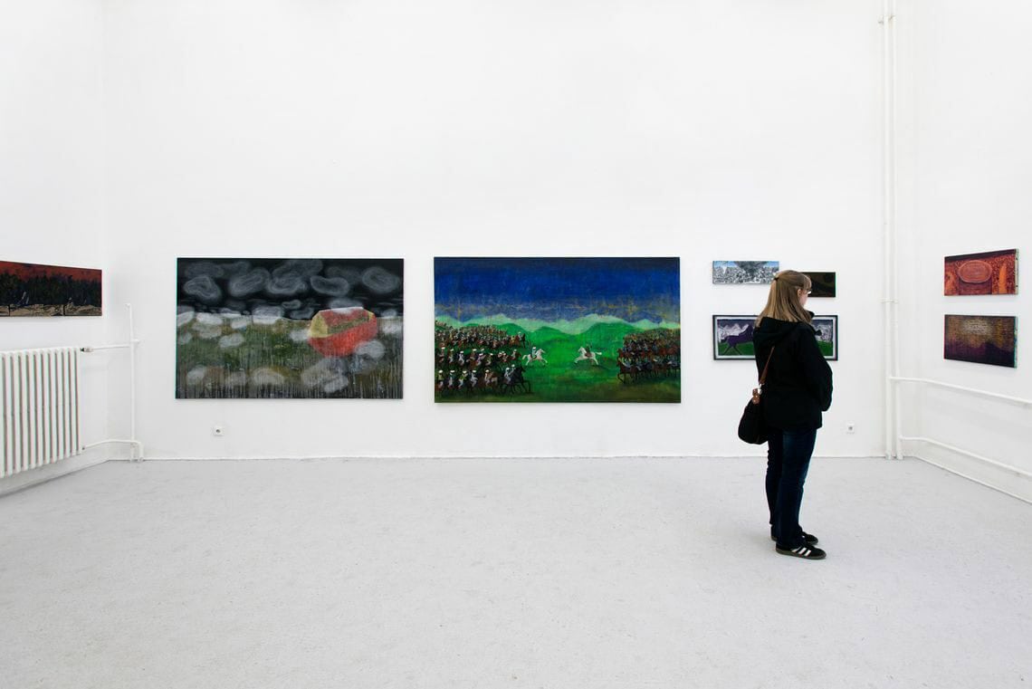 Foto: Ein Raum mit weißen Wänden und grauem Boden .An den Wänden hängen, auf ähnlicher Höhe, mehrere Leinwände und gerahmte Objekte. im Raum steht ein Mensch.