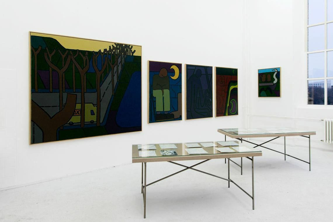 Foto: Ein Raum mit weißen Wänden, grauem Boden und einem großen Fenster. An den Wänden hängen mehrere Leinwände in Klein- und Mittelformaten. Im Raum stehen außerdem zwei Tische, auf denen Glasplatten liegen. Unter den Glasplatten liegen Papiere.