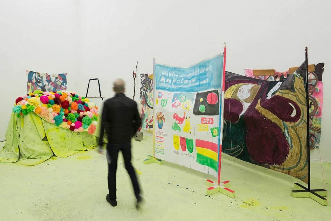 Foto: Ein räum mit weißen Wänden und grünem Boden. Im Raum befinden sich viele Dinge, die nah beieinander stehen. Darunter ein Haufen aus Material und bemalte Textilien, die zwischen Stöcken aufgespannt sind. Durch den Raum bewegt sich ein Mensch.