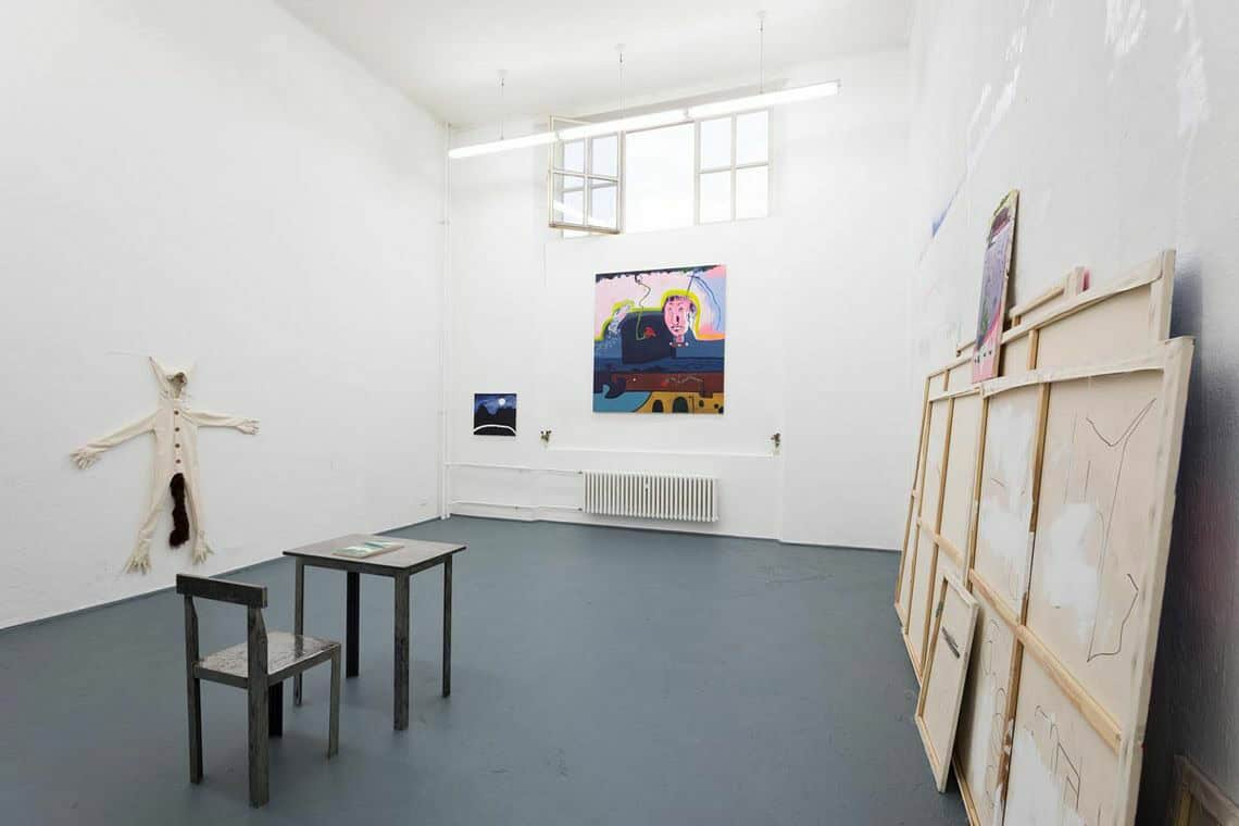 Foto: Ein Raum mit weißen Wänden und grauem Boden. Teile des Fensters sind mit einer weißen Platte bedeckt. Rechts an der Wand lehnt eine Gruppe von Leinwänden. An einer anderen Wand hängen zwei Leinwände im Klein- und Großformat. Links an der Wand hängt ein Objekt. Im Raum stehen ein Stuhl und ein Kleiner Tisch, die aus dem gleichen material gefertigt sind. Auf dem Tisch liegt ein Heft.