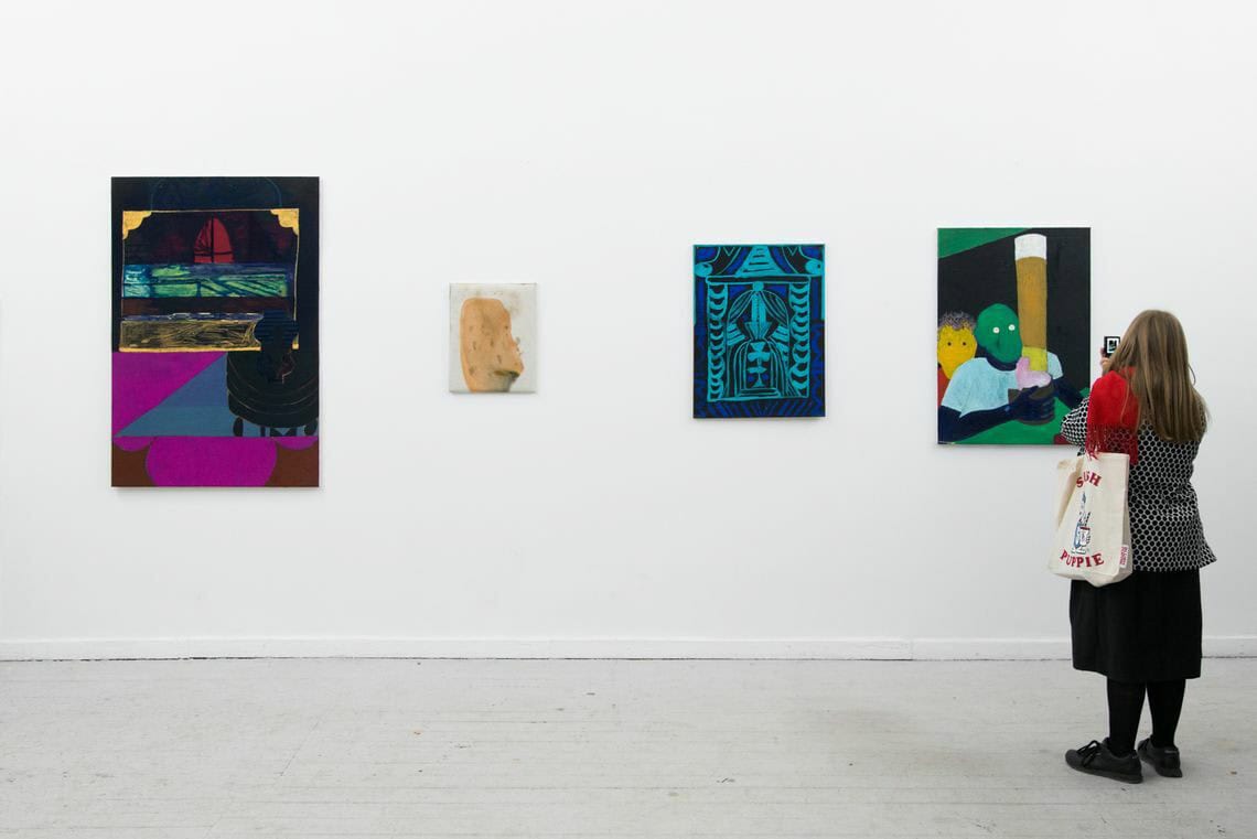 Foto: Ein Raum mit weißer Wand und grauem Boden. An der Wand hängen, auf gleicher Mittellinie, Leinwandarbeiten im Klein- und Mittelformat. Im Raum steht eine Person.