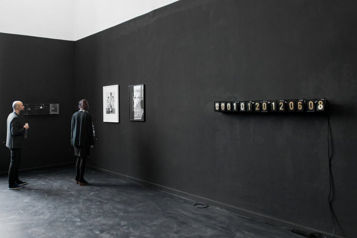 Foto: Ein Raum mit schwarzer Wand und schwarzem Boden. Der obere Teil der Wand ist weiß gestrichen. Auf der schwarzen Fläche hängen mehre flache Objekte auf unterschiedlichen Materialien. Im Raum stehen zwei Menschen.