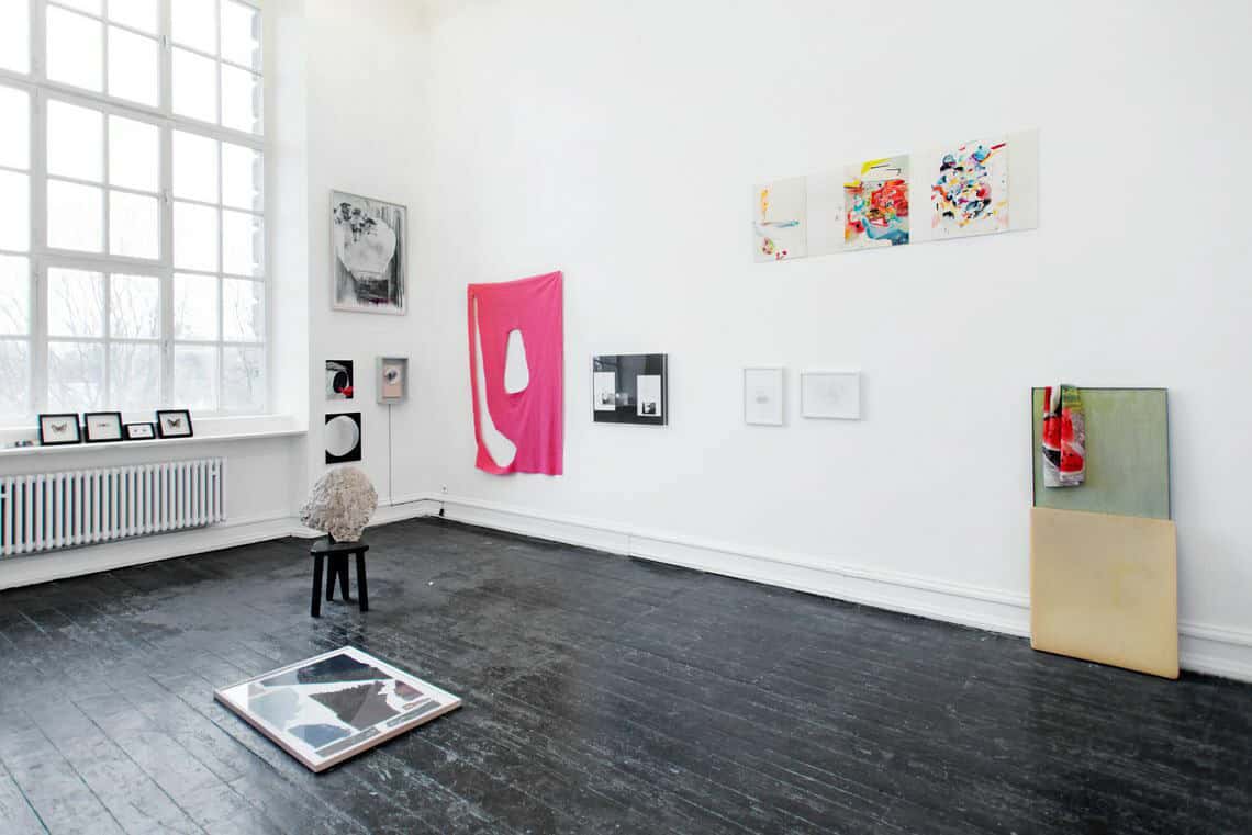 Foto: Ein Raum mit weißen Wänden, schwarzem Dielenboden und einem großen Fenster. An den Wänden und auf der Fensterbank befinden sich mehrere flache Objekte, einige von ihnen sind gerahmte, andere nicht. Auf dem Boden stehen und liegen ebenfalls Objekte.
