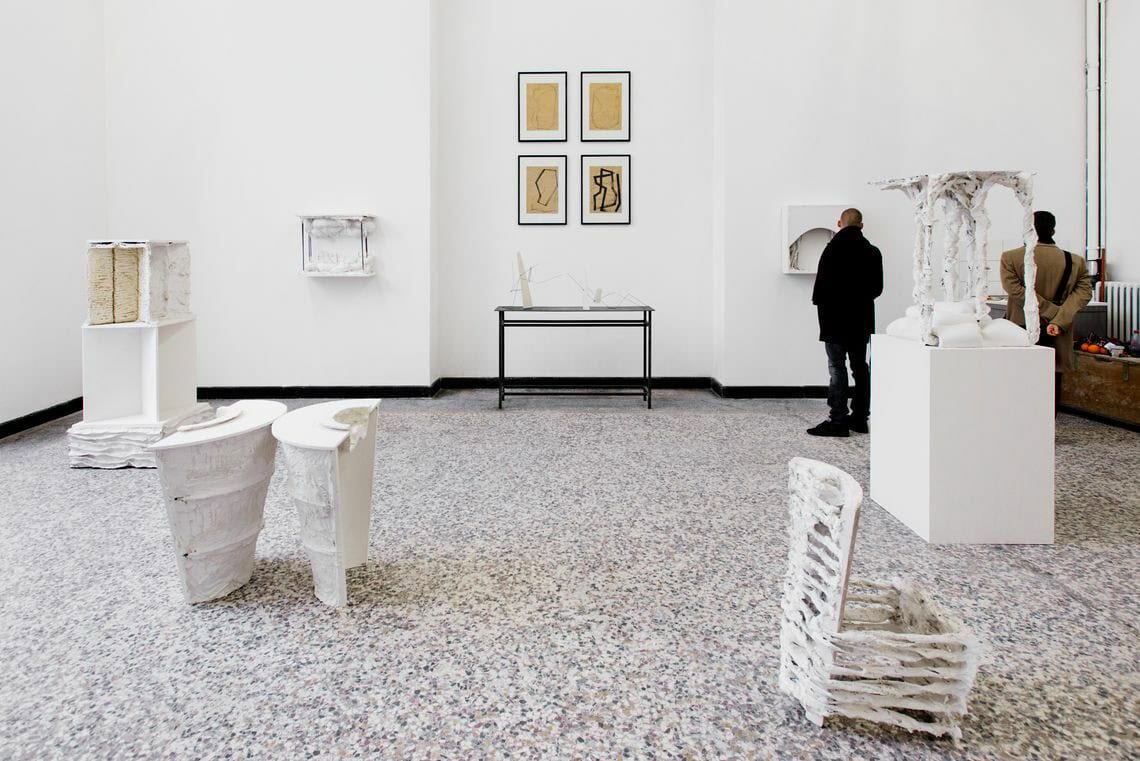 Foto: Ein Raum mit weißen Wänden und mehrfarbigem Steinboden. An den Wänden befinden sich mehrere Objekte, einige von ihnen sind plastisch, andere flach und gerahmt. Im Raum befinden sich mehrere große Skulpturen aus unterschiedlichen Materialien. Sie stehen auf dem Boden.