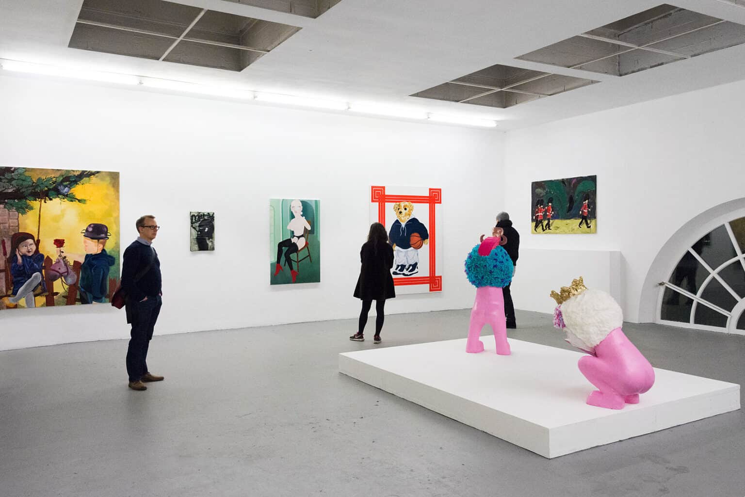 Foto: Ein Raum mit weißen Wänden, grauem Boden und einem kleinen Bogenfenster. An den Wänden hängen Leinwände im Klein-, Mittel- und Großformat. Im Raum steht ein flächiges, quadratisches Podest, auf dem sich zwei Skulpturen befinden. Durch den Raum bewegen sich Menschen.