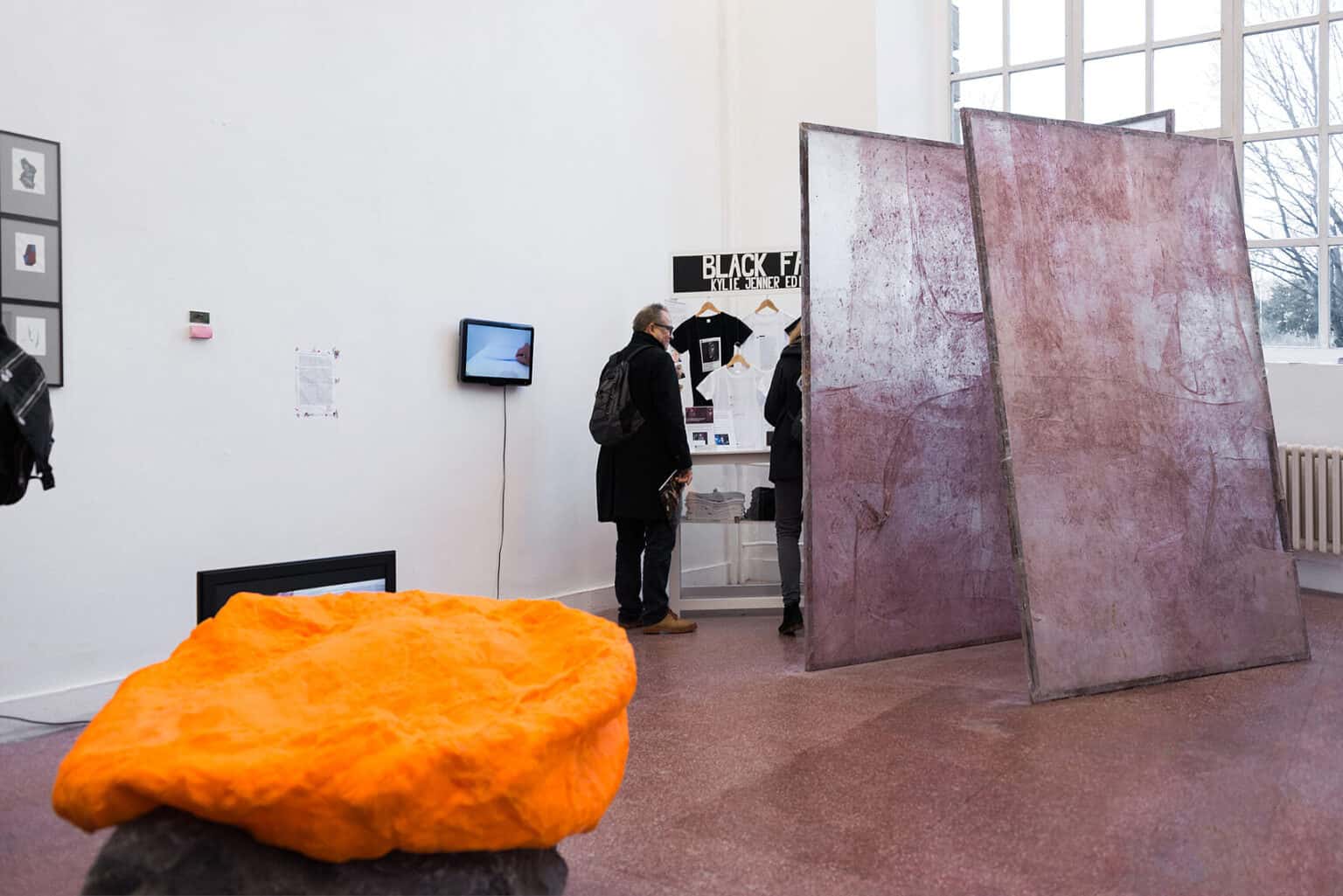 Foto: Ein Raum mit weißen Wänden, rotem Steinboden und einem großen Fenster. An der Wand des Raumes hängen unterschiedliche Dinge, darunter Zettel, gerahmte Objekte und Bildschirme. Im gesamten Raum verteilt befinden sich Skulpturen auf unterschiedlichen Materialien in unterschiedlicher Größe. im Raum befinden sich zwei Menschen.