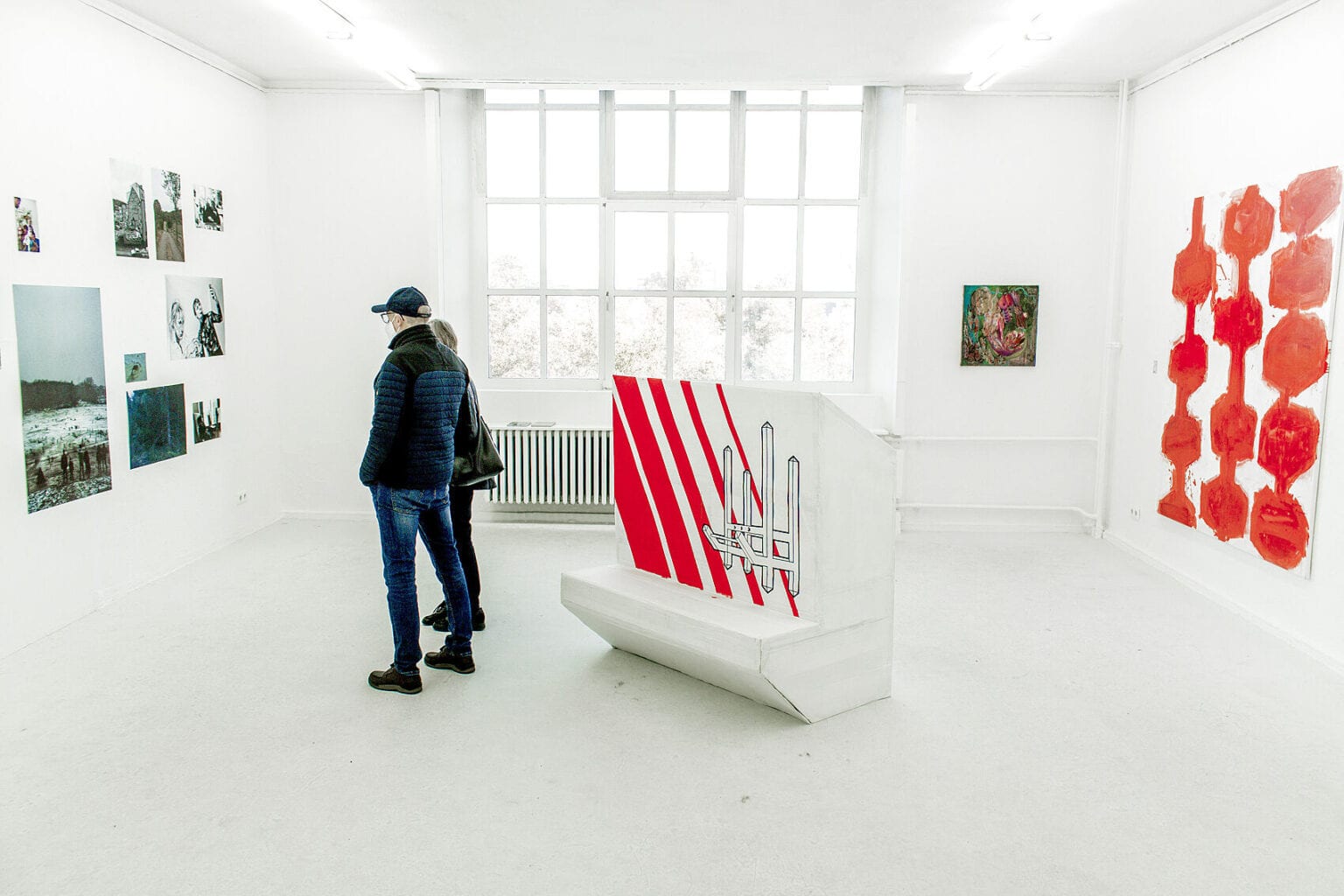 Foto: Ein Raum mit weißen Wänden, hellem Boden und einem Fenster. An den Wänden befinden sich Leinwandarbeiten im Klein- und Großformat und eine Gruppe von Flachen Objekten. Im Raum stehen zwei menschen und ein verschachteltes Objekt.