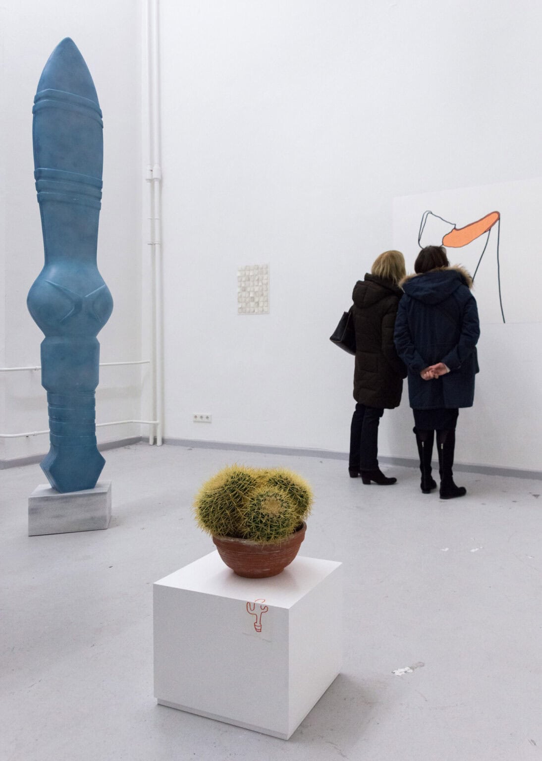 Foto: Ein Raum mit weißen Wänden und grauem Boden. An der Wand hängen zwei flache Objekte. Vor der Wand stehen zwei Menschen. Auf dem Boden stehen kleine und große Skulpturen auf Sockeln.