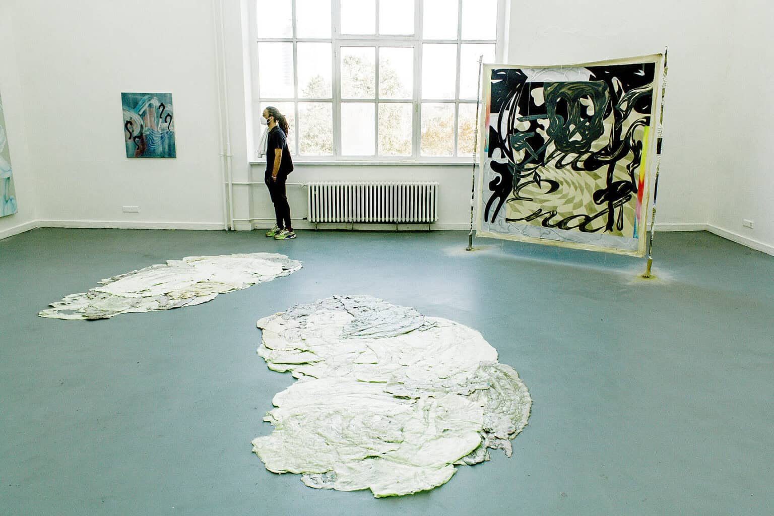 Foto: Ein Raum mit weißen Wänden, grauem Boden und einem großen Fenster. Auf dem Boden liegen zwei flache, flächige Objekte. Daneben steht ein auf zwei Stelzen befestigter Banner. Vor dem Fenster steht ein Mensch. An der Wand hält eine mittelformatige Leinwandarbeit.