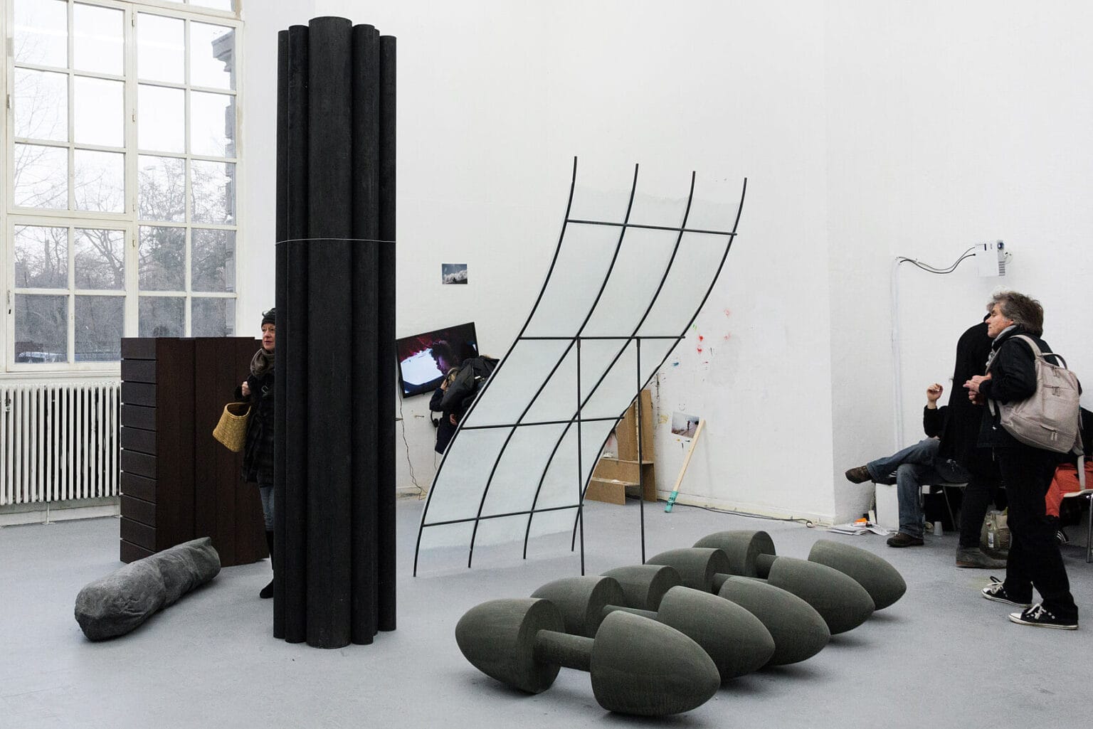 Foto: Ein Raum mit weißen Wänden, grauem Boden und einem großen Fenster. Im Raum befinden sich viele große Skulpturen, die nah beieinander auf dem Boden stehen. An der Wand hängen und lehnen Objekte aus unterschiedlichen Materialien, darunter ein Bildschirm, der nicht gerade hängt. Durch den Raum bewegen sich mehrere Menschen.