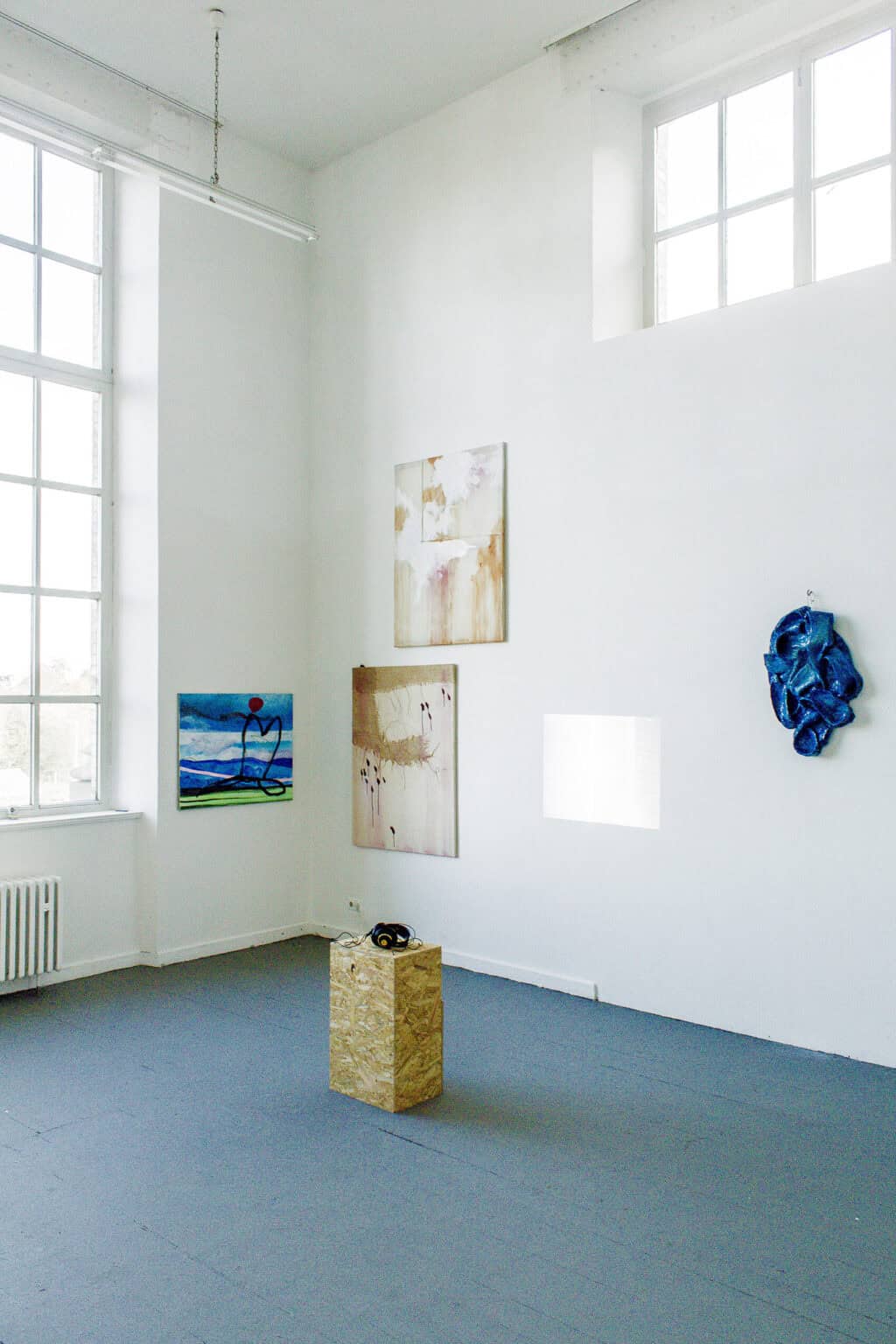 Foto: Ecke eines Raumes mit weißen Wänden und grauem Dielenboden. Im Raum befindet sich ein Sockel, auf dem ein Kopfhörer liegt. An den Wänden hängen, auf unterschiedlicher Höhe, Objekte und Leinwandarbeiten.