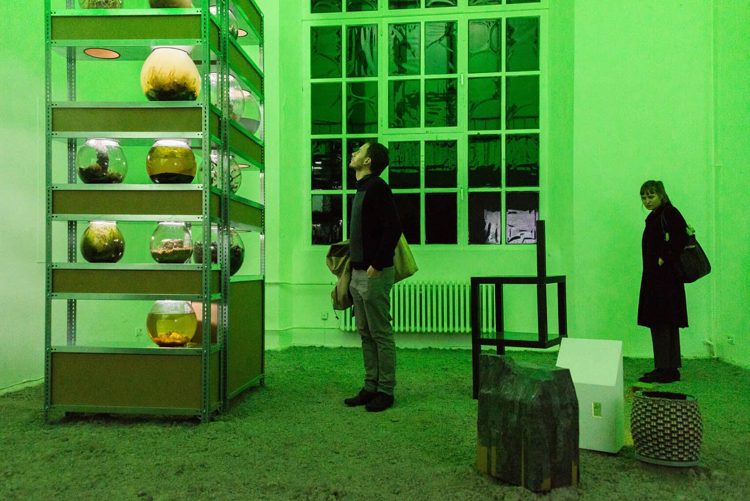 Foto: Ein Raum mit weißen Wänden und großem Fenster. Der Boden ist mit einem erdigen oder sandigen Material bedeckt. Im Raum stehen Objekte auf dem Boden und eine große Regalkonstruktion, in der sich gefüllte, runde Behältnisse befinden. Der gesamte Raum ist in grünes Licht getaucht. Im Raum befinden sich zwei Personen.