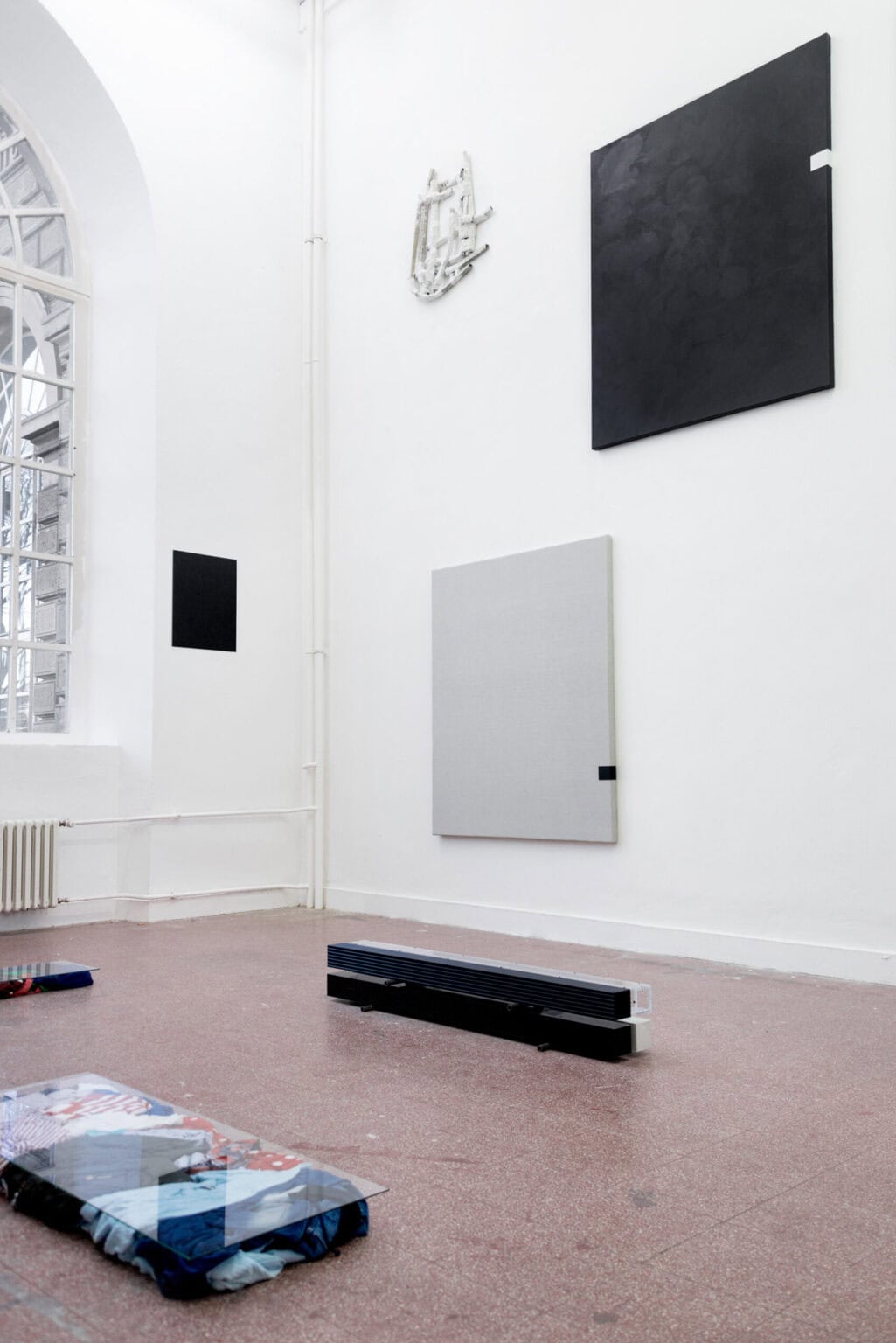 Foto: Ein Raum mit weißen Wänden und rotem Steinboden. An der Wand hängen, über die gesamt Höhe der Wand verteilt, flache Objekte. Auf dem Boden liegen mehrere Skulpturen.