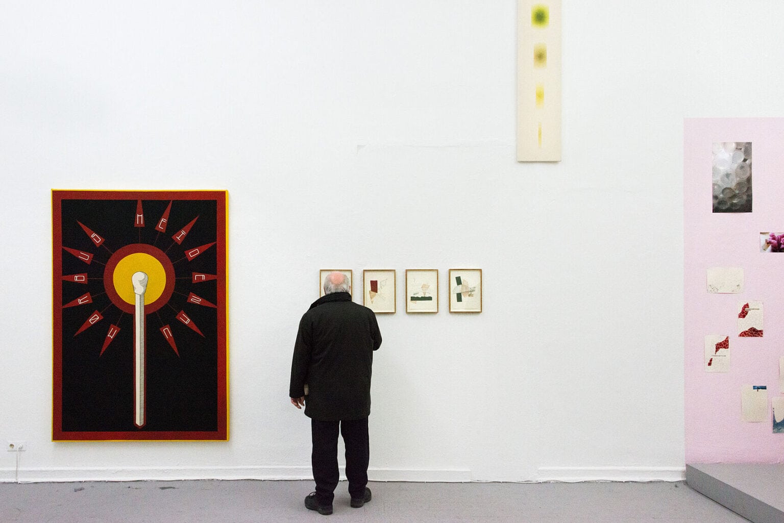 Foto: Ein Raum mit weißer Wand und grauem Boden. rechts auf der Wand befindet sich ein rosafarbener Streifen. Auf dem Boden vor dem Streifen liegt ein graues Podest. An der Wand hängen flache Objekte in unterschiedlichen Formaten auf unterschiedlicher Höhe. Einige der Objekte sind gerahmt. Im Raum steht ein Mensch.