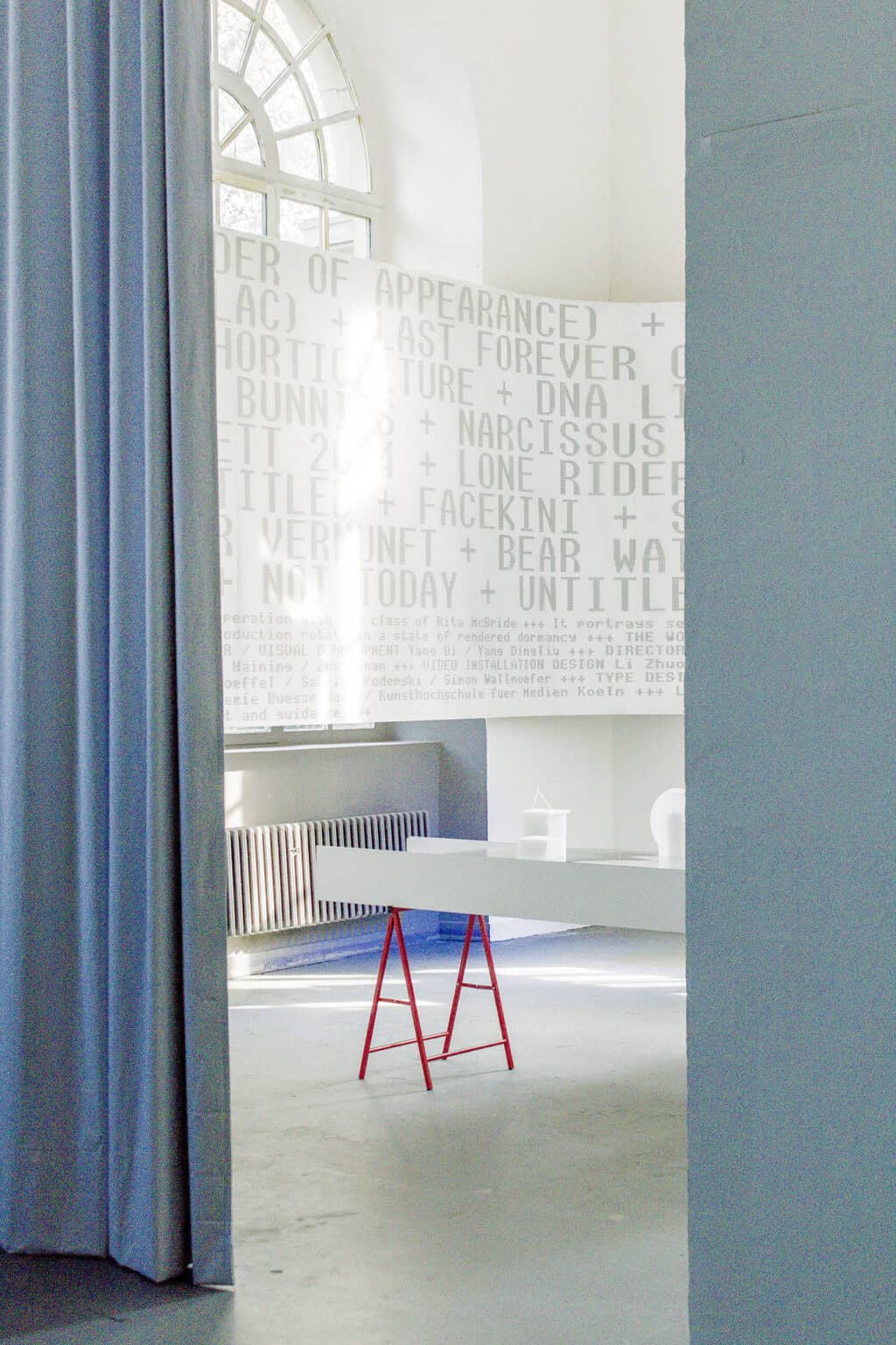 Foto: Ein Raum mit weißen Wänden, grauem Boden und einem großen Fenster. Im Raum steht ein roter Bock, auf dem ein Kasten liegt, auf dem sich Objekte befinden. Über die Wand und einen Teil des Fenster erstreckt sich ein weißer, breiter Banner, der mit grauer Schrift versehen ist.