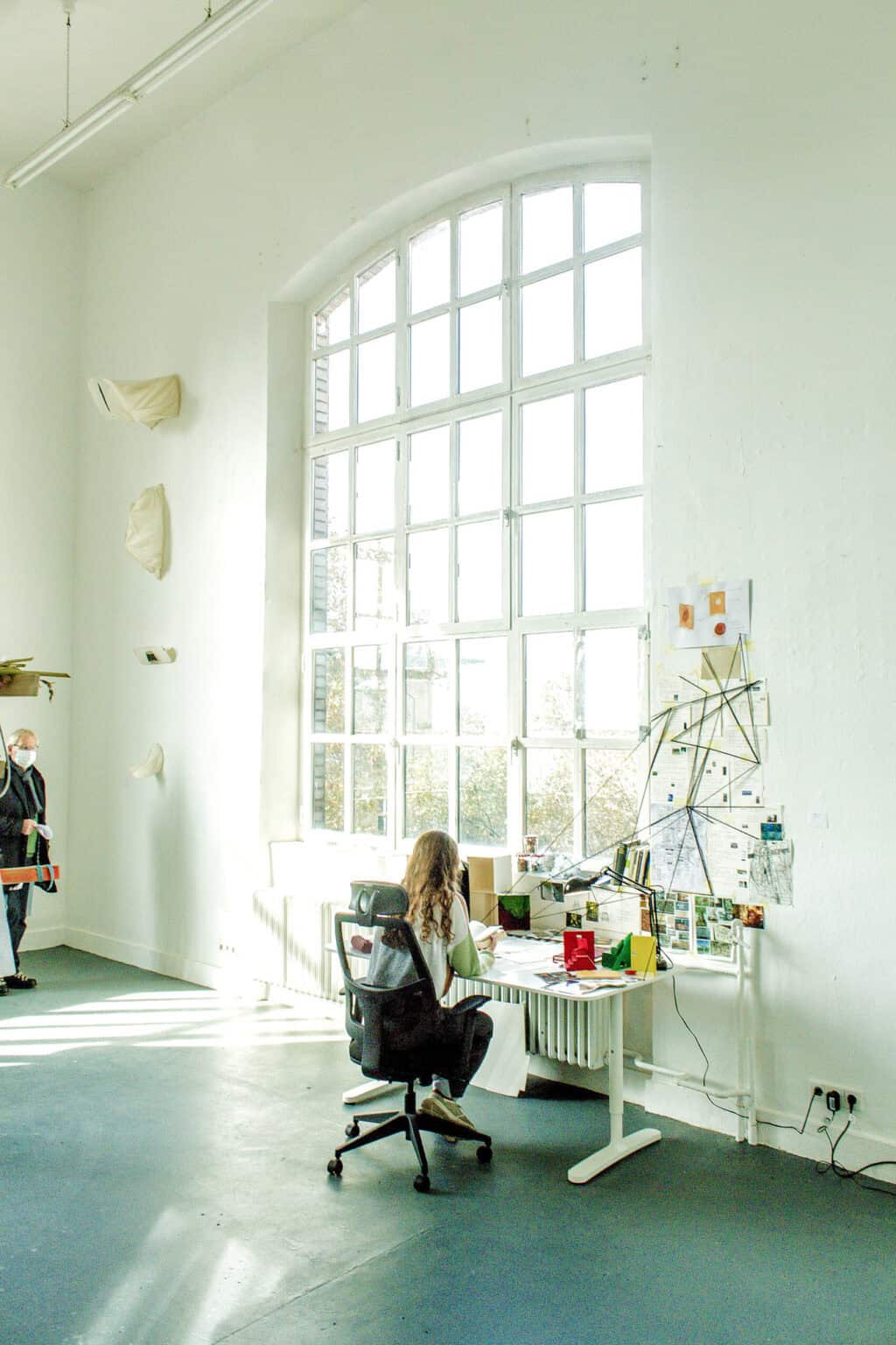 Foto: Ein Raum mit weißen Wänden, grauem Boden und einem großen Fenster. An Der Wand hängen, in einer Reihe untereinander, Objekte. Vor dem Fenster ist ein Schreibtisch aufgebaut, vor dem auf einem Stuhl eine Person sitzt. Der Schreibtisch ist vollgestellt mit Zetteln und Objekten. An einem Wandstück vor dem Schreibtisch hängen Zettel und Abbildungen, auf denen in kreuzförmigen Muster Schnüre verlaufen. Die Schüre sind zwischen der Wand und dem Schreibtisch gespannt.