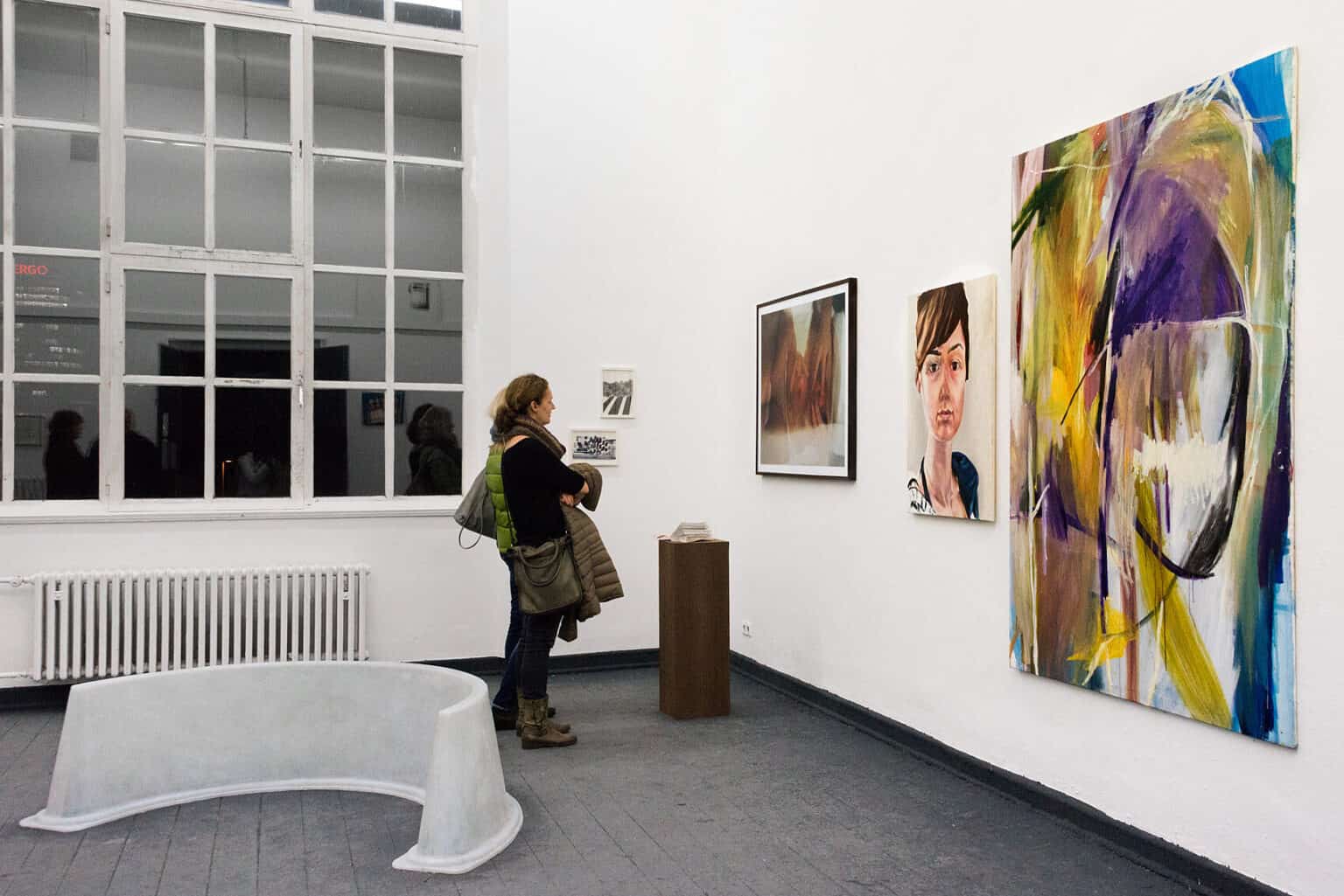 Foto: Ein Raum mit weißen Wänden, grauem Dielenboden und einem großen Fenster. An der Wand hängen gerahmte und ungerahmte Objekte im Klein-, Mittel- und Großformat. Im Raum stehen Skulpturen direkt auf dem Boden und auf Sockeln. Im Raum stehen zwei Menschen.