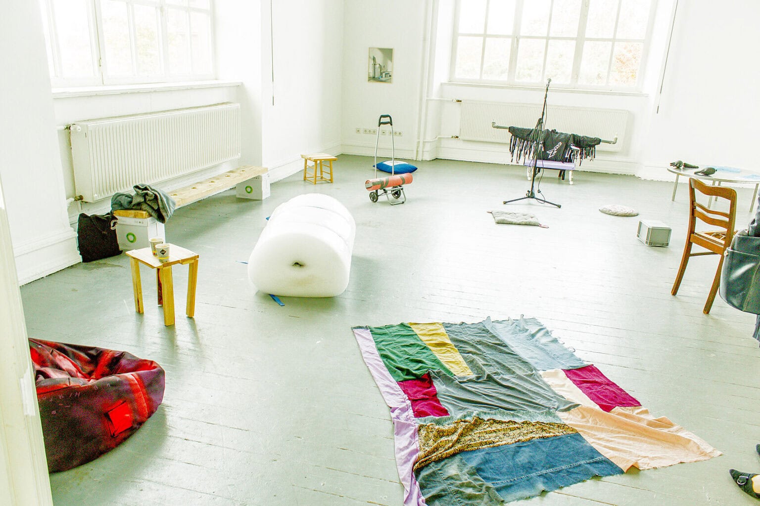 Foto: Ein Raum mit weißen Wänden, grauem Dielenboden und zwei goßen Fenstern. Auf dem Boden des Raumes verteilt liegen und stehen eine Vielzahl an Objekten, darunter Textilien, Luftpolsterfolie, Tische, Stühle, Sackkarren, Sessel, Boxe, Stative usw. An der Wand hängt eine kleinformatige, flache Arbeit.
