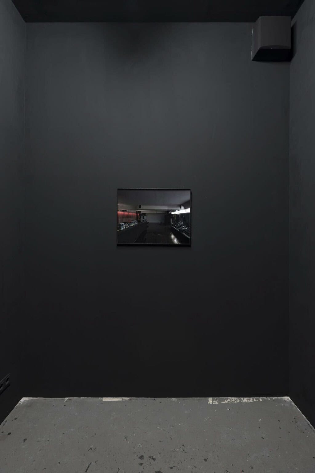 Foto: Ein schmaler Raum mit schwarzer Wand und grauem Boden. An der Wand befindet sich ein flaches, gerahmtes Objekt.