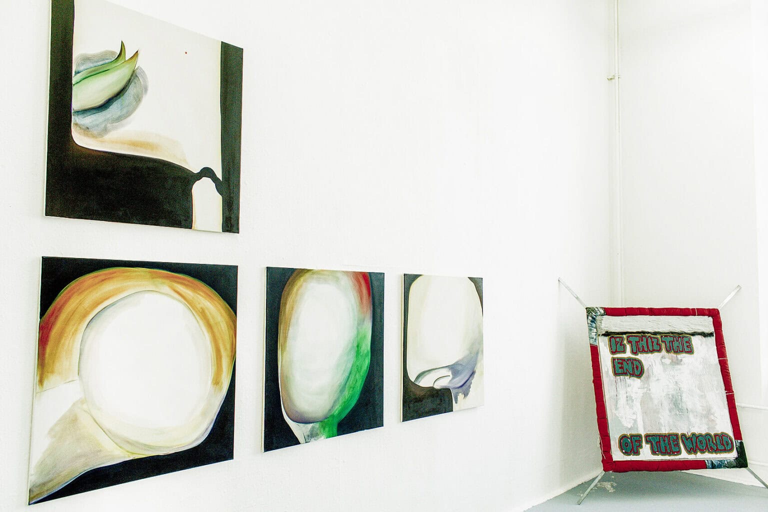 Foto: Eine Raumecke mit weißen Wänden und grauem Boden. An der Wand hängen, in zwei reihen, quadratische Leinwände. In der Ecke befindet sich ein quadratisches Objekt, dass auf dem Boden steht und an der Wand lehnt.