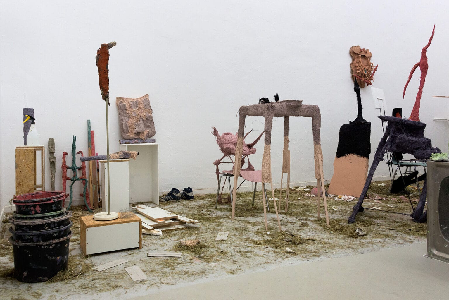 Foto: Ein Raum mit weißen Wänden und grauem Boden. Auf dem Boden des Raumes befindet sich getrocknetes Gras und ein Arrangement aus vielen unterschiedlichen Objekten, darunter Eimer, Sockel, Schuhe und Skulpturen, die nah beieinander stehen.