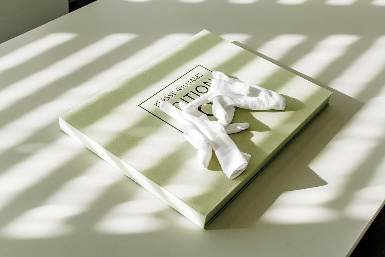 Foto:Eine Flache, quadratische Box auf einer hellen Tischplatte. Auf der Box der Schriftzug "Klasse Williams" und ein weißes, verdrehtes paar Handschuhe.